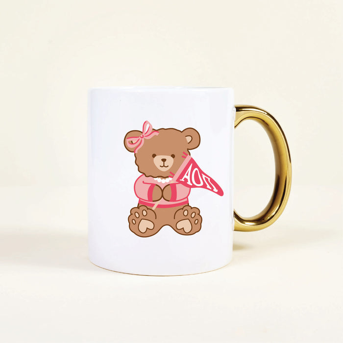 Enjoy your morning coffee or tea with The Darling Effect Alpha Omicron Pi 11 oz Cozy Cute Ceramic Mug, featuring a sweet bear in a pink bow and shirt holding an “AOII” pennant and accented with a gold handle—perfect for any sorority member.