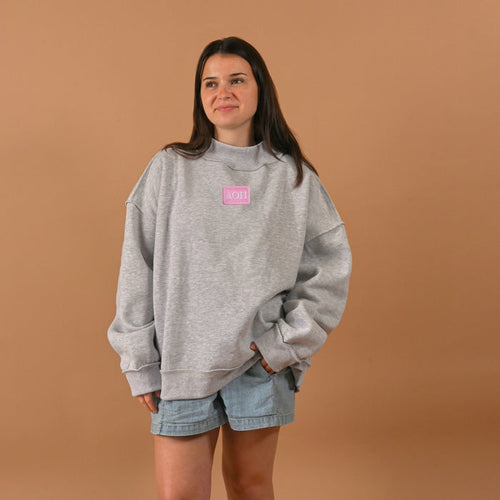 A woman stands against a plain brown background wearing The Darling Effect’s Alpha Omicron Pi Letter Patch Mockneck Sweatshirt (S/M) in gray and light blue denim shorts, her hands in her pockets and a relaxed expression.