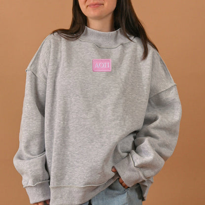 A person with long brown hair, whose face is partially visible, is wearing The Darling Effect’s Alpha Omicron Pi Letter Patch Mockneck Sweatshirt in size S/M—an oversized gray sorority sweatshirt featuring a pink patch with Greek letters. The background is solid beige.