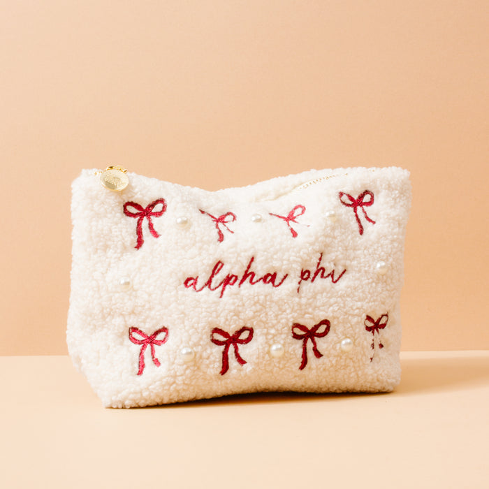 The Darling Effect’s Alpha Phi Bows & Pearls Teddy Pouch is a cream sherpa pouch with red embroidered bows, pearl accents, and "alpha phi" stitched on the front—a perfect sorority gift—shown against a peach background.