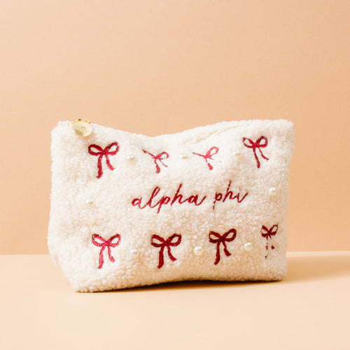 The Darling Effect’s Alpha Phi Bows & Pearls Teddy Pouch is a cream sherpa pouch with red embroidered bows, pearl accents, and "alpha phi" stitched on the front—a perfect sorority gift—shown against a peach background.
