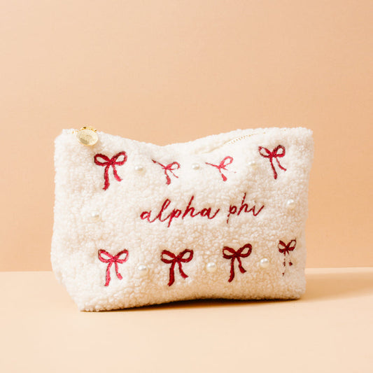 The Darling Effect’s Alpha Phi Bows & Pearls Teddy Pouch is a cream sherpa pouch with red embroidered bows, pearl accents, and "alpha phi" stitched on the front—a perfect sorority gift—shown against a peach background.