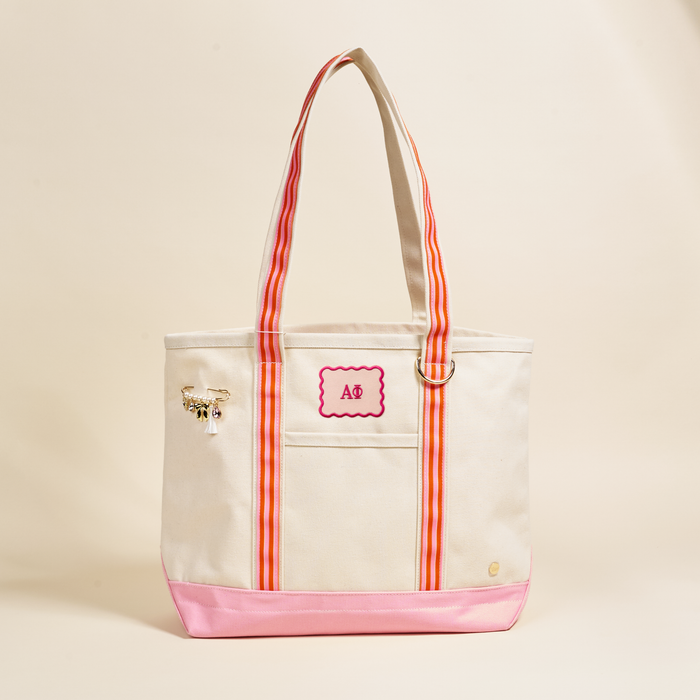 The Darling Effect’s Alpha Phi Canvas Tote Bag features pink trim, striped handles, a gold pin, a scalloped “AB” patch, and a gold ring on the beige front—perfect as an everyday or sorority bag. Plain light beige background.