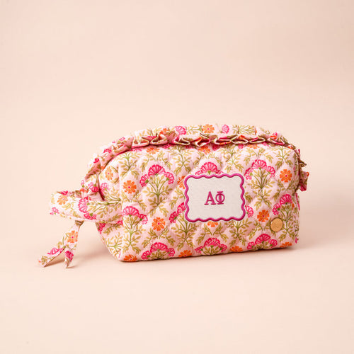 The Darling Effect’s Alpha Phi Essentials Quilted Cosmetic Pouch features pink, orange, and green floral petals, a white label with pink “ΑΦ” Greek letters, and a knotted fabric zipper pull—ideal for carrying your sorority must-haves.