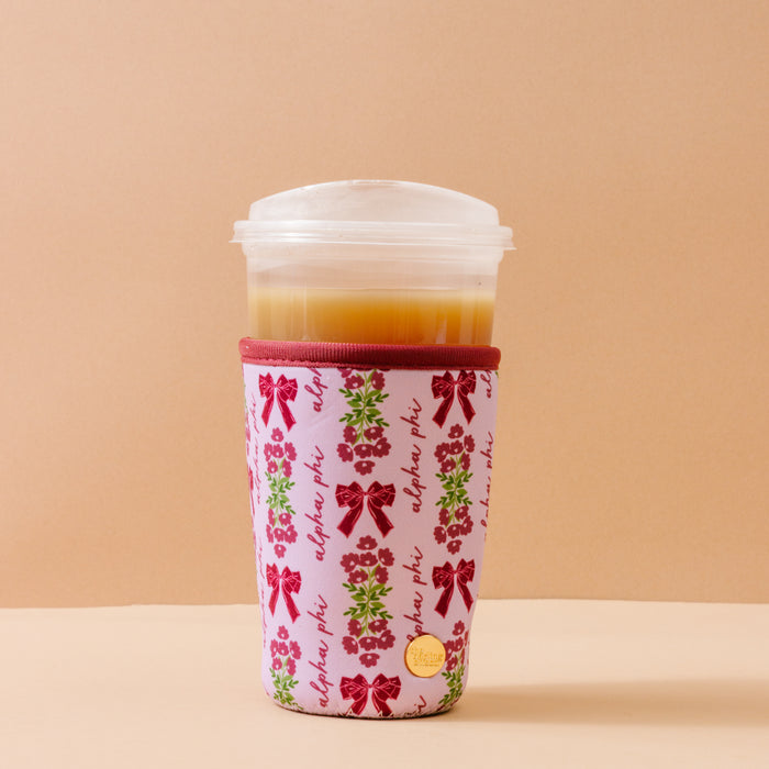 A plastic cup of iced coffee with a lid is wrapped in The Darling Effect's Alpha Phi Large Sip Savvy Drink Sleeve, featuring red bows, green wreaths, and "alpha phi" in red script—an ideal sorority gift against a beige background.
