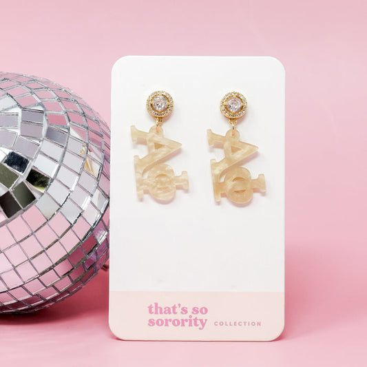 Alpha Phi Letter Gold Acrylic Earrings by The Darling Effect, featuring gold and pearl details on a white "that's so sorority collection" card—perfect for sisterhood events or bid-day baskets, shown with a disco ball and pink background.