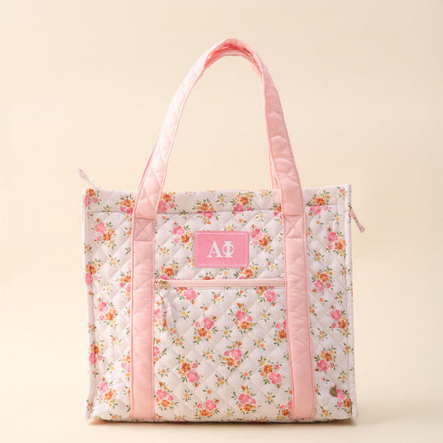 The Darling Effect's Alpha Phi The Everyday Quilted Tote Bag features a floral quilted design, light pink handles and trim, a front pocket with white Greek letters on a pink patch—ideal for displaying your sorority pride in style.