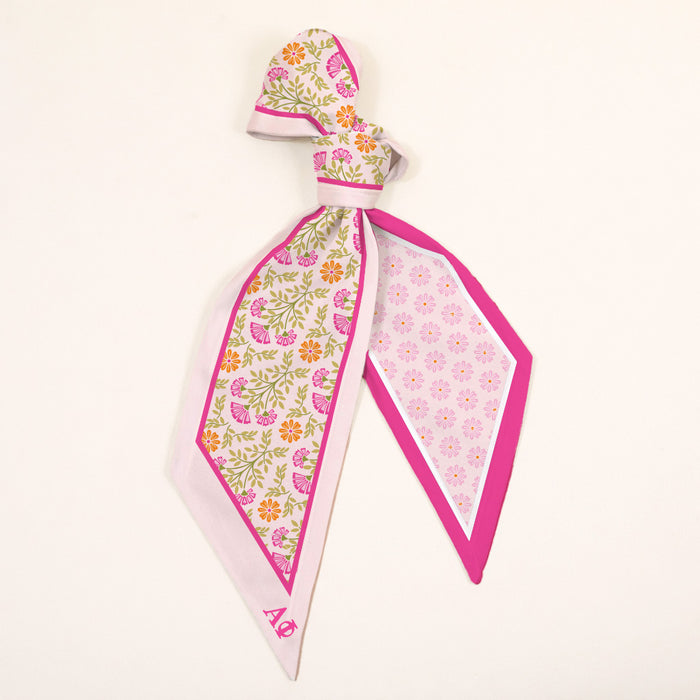 The Darling Effect’s Alpha Phi Twirl & Tie Scarf Accessory features pink, yellow, and orange floral patterns with a pink border and Alpha Phi initials in one corner, tied elegantly against a plain white background.