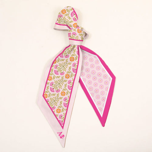 The Darling Effect’s Alpha Phi Twirl & Tie Scarf Accessory features pink, yellow, and orange floral patterns with a pink border and Alpha Phi initials in one corner, tied elegantly against a plain white background.