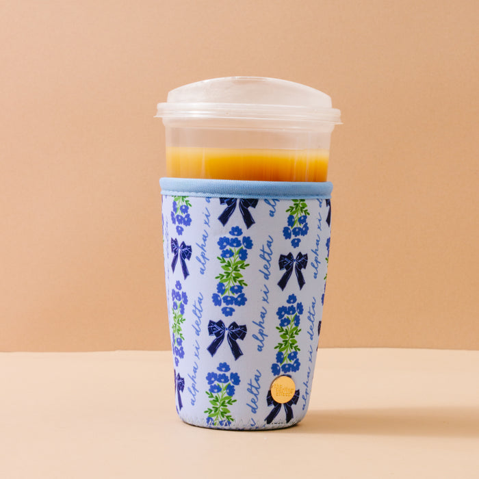 A plastic cup of orange juice with a clear lid is held in The Darling Effect’s Alpha Xi Delta Large Sip Savvy Drink Sleeve, featuring blue bows and “alpha xi delta” on beige—perfect as a sorority gift.
