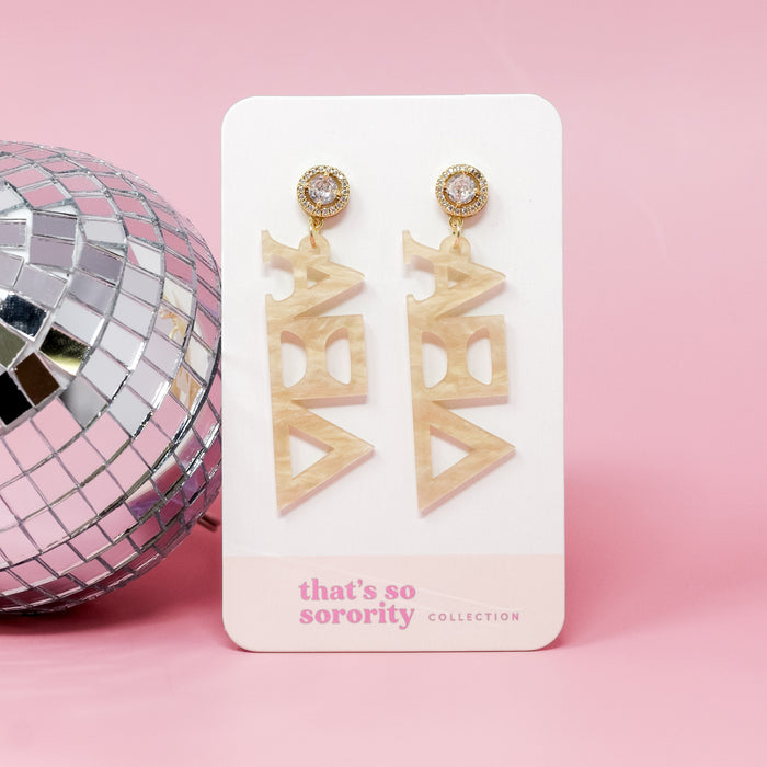 A pair of Alpha Xi Delta Letter Gold Acrylic Earrings by The Darling Effect hang from a white card, featuring a pearlescent finish and clear rhinestones—perfect for sisterhood events or as a stylish bid-day basket gift.