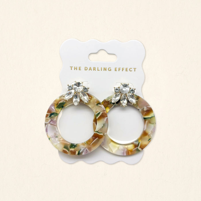 The Amber Drift Glow Getter Earrings by The Darling Effect feature glossy marbled resin, clear crystals, and gold hoops on a white scalloped card with The Darling Effect logo.