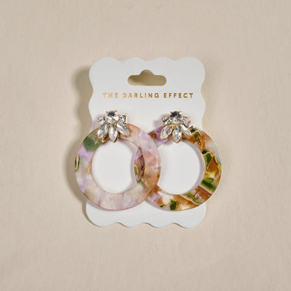 Amber Drift Glow Getter Earrings by The Darling Effect: round, multicolored marbled earrings with rhinestone flower-shaped studs, shown on a white THE DARLING EFFECT card against a beige background.