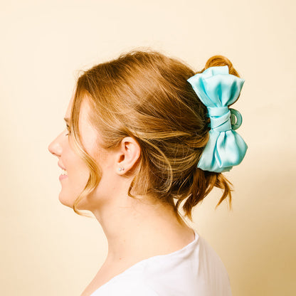 A woman with light brown hair smiles in profile, wearing a white shirt and securing her updo with the-darling-effect Aqua Satin Bow Claw Clip against a beige background.