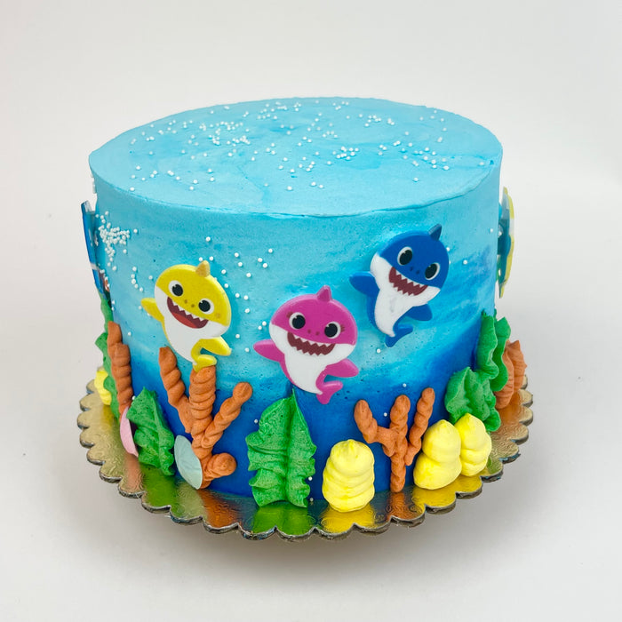 A decorated cake with Baby Shark characters and sea life accents in blue, pink, yellow, and green colors.