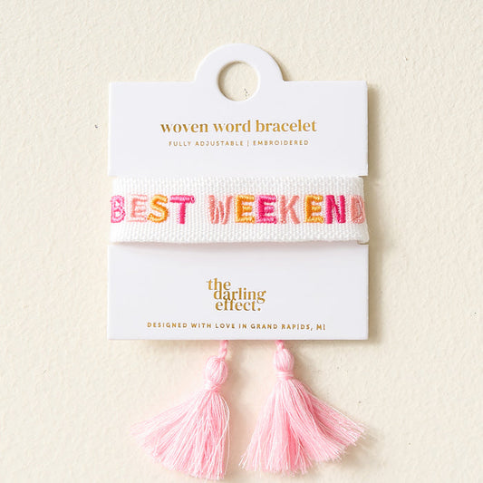 The Darling Effect Best Weekend Woven Word Bracelet features "BEST WEEKEND" embroidered in colorful letters on lightweight white woven material, with two pink tassels, displayed on a card reading "Woven Word Bracelet" and "The Darling Effect.