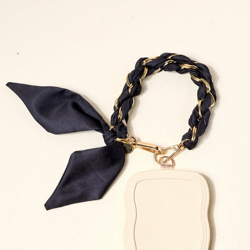 A stylish black phone scarf from The Darling Effect featuring a braided wrist strap with gold-tone chain accents and a bow, attached to an ID holder—a chic accessory.
