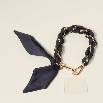The Darling Effect's Black Call Me Later Phone Scarf features a gold-tone chain interwoven with black fabric, a decorative black bow, and a gold charm—a chic accessory for any occasion, displayed on a plain beige background.