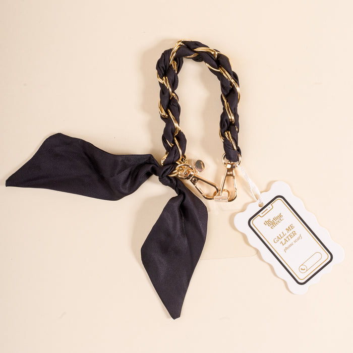 The Darling Effect’s Black Call Me Later Phone Scarf features black fabric braided with a gold-tone chain, a clasp, and a white “Call Me Later” tag, all displayed on a light beige background.