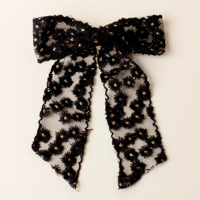 The-darling-effect Black Ditzy Daisies Hair Bow features sheer lace with floral embroidery, gold metallic details, long trailing tails, scalloped edges, and a secure gold barrette clip, shown on a white background.