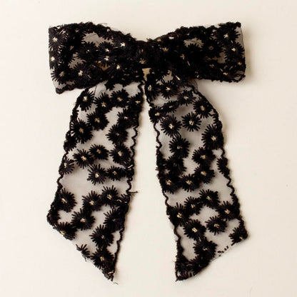 The-darling-effect Black Ditzy Daisies Hair Bow features sheer lace with floral embroidery, gold metallic details, long trailing tails, scalloped edges, and a secure gold barrette clip, shown on a white background.