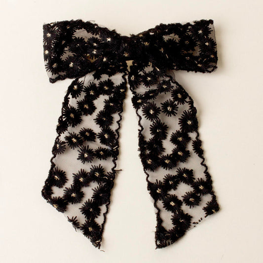 The-darling-effect Black Ditzy Daisies Hair Bow features sheer lace with floral embroidery, gold metallic details, long trailing tails, scalloped edges, and a secure gold barrette clip, shown on a white background.