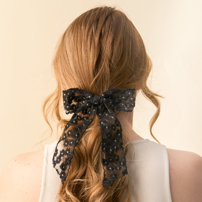 A woman with long, wavy light brown hair is seen from behind in a white sleeveless top, her hair styled with the-darling-effect's Black Ditzy Daisies Hair Bow, adding an elegant touch to her look.