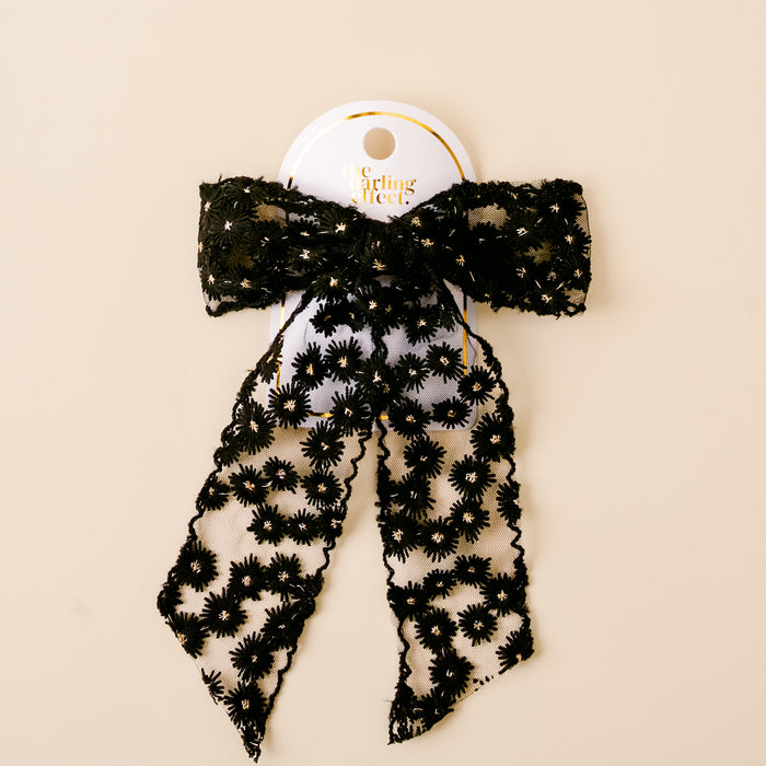 The Black Ditzy Daisies Hair Bow by the-darling-effect features black lace with floral patterns, attached to a white card with gold text and displayed against a beige background.