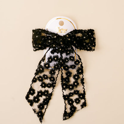 The Black Ditzy Daisies Hair Bow by the-darling-effect features black lace with floral patterns, attached to a white card with gold text and displayed against a beige background.