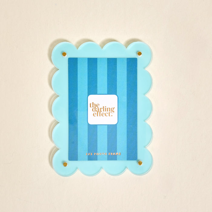 The Darling Effect Blue Mini Acrylic Picture Frame Magnet features scalloped edges, vertical blue stripes, and a white brand label. It perfectly fits 2x3 Polaroids and is ideal for displaying your favorite photos on the fridge.