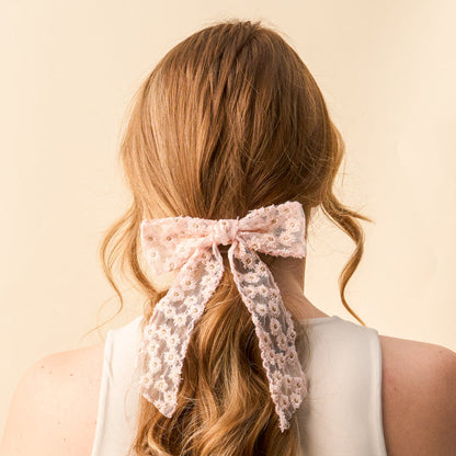 A woman with wavy, light brown hair wears a white top and ties her hair with the Blush Ditzy Daisies Hair Bow from the-darling-effect, featuring a large sheer floral design. The background is soft beige.