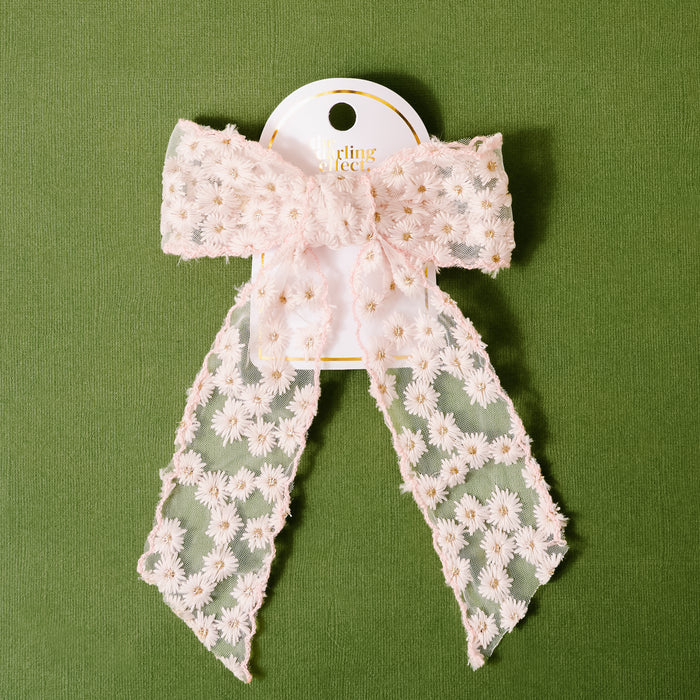The Blush Ditzy Daisies Hair Bow by the-darling-effect, a sheer light pink bow with embroidered white flowers, is attached to a white card and showcased on a textured green background.