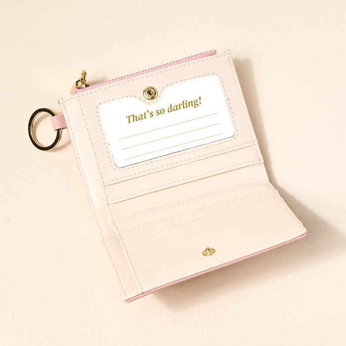 The Darling Effect’s Blush Luxe Pocket Wallet, a pale pink vegan leather accessory, features card slots, a clear ID window with a “That’s so darling!” insert, and a gold keyring, shown open on a light surface.