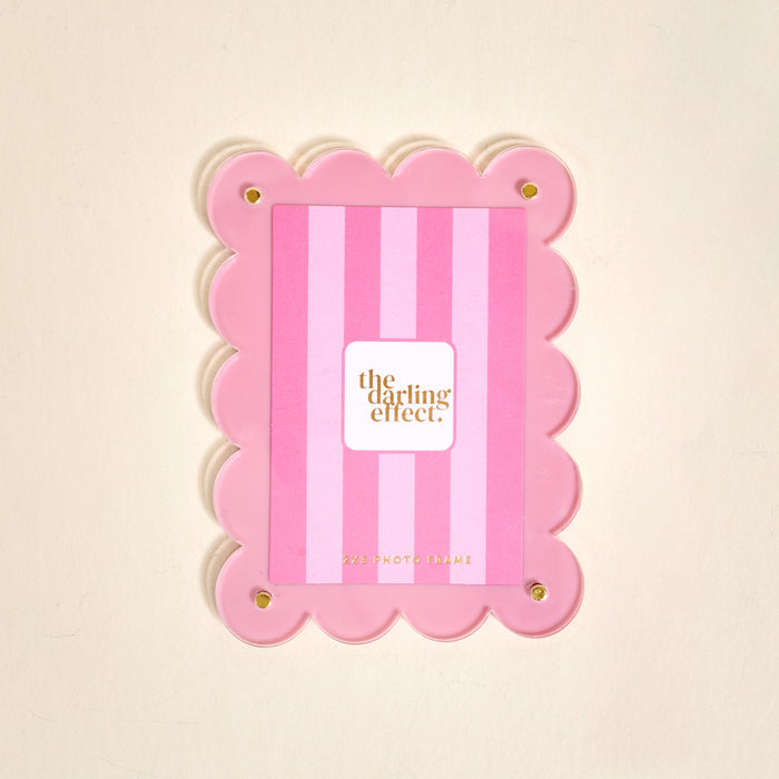 The Darling Effect’s Blush Mini Acrylic Picture Frame Magnet features scalloped edges, vertical blush stripes, and four gold corner bolts—perfect for displaying polaroid photos. The center displays a card with the brand name.