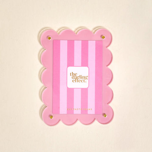 The Darling Effect’s Blush Mini Acrylic Picture Frame Magnet features scalloped edges, vertical blush stripes, and four gold corner bolts—perfect for displaying polaroid photos. The center displays a card with the brand name.