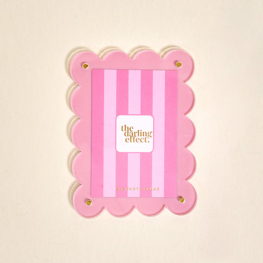 The Darling Effect’s Blush Mini Acrylic Picture Frame Magnet features scalloped edges, vertical blush stripes, and four gold corner bolts—perfect for displaying polaroid photos. The center displays a card with the brand name.