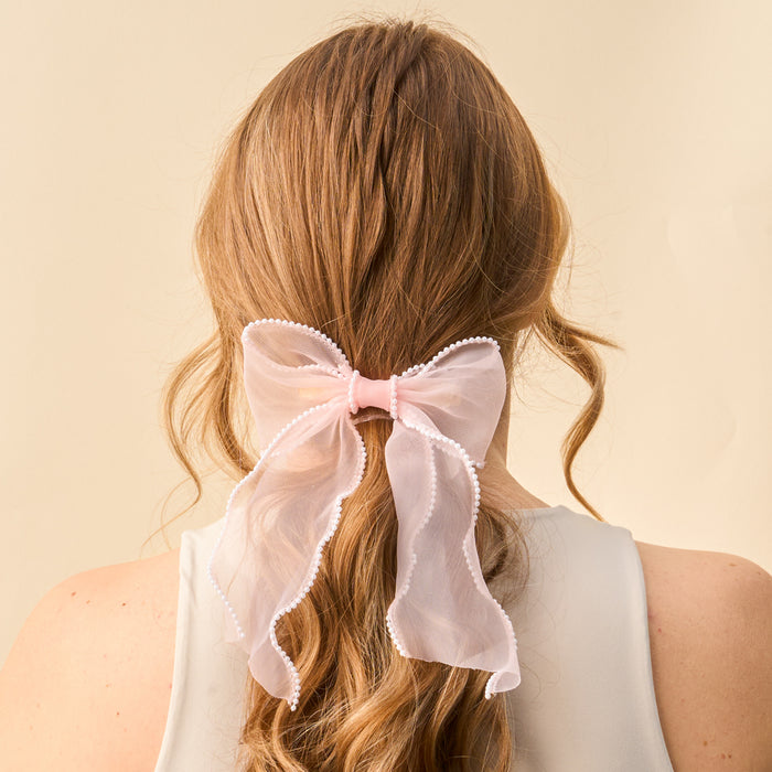 The Blush Sheer Pearl Hair Bow from the-darling-effect features a large, sheer pink bow adorned with white pearls and ribbon accents—a charming hair accessory for long, wavy blonde styles.
