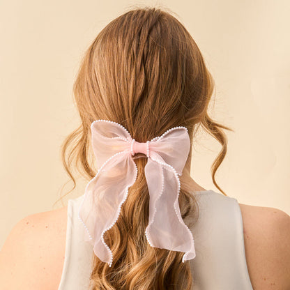 The Blush Sheer Pearl Hair Bow from the-darling-effect features a large, sheer pink bow adorned with white pearls and ribbon accents—a charming hair accessory for long, wavy blonde styles.
