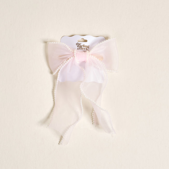 The Blush Sheer Pearl Hair Bow by the-darling-effect features light pink sheer fabric, elegant pearl trim, and long flowing tails, presented on a branded white card.