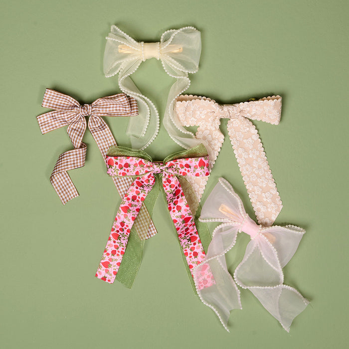 Five decorative hair bows on a green background, including the Blush Sheer Pearl Hair Bow by the-darling-effect, plus a brown gingham bow, pink strawberry print with green ribbon, cream lace, and a pink sheer pearl bow—charming accessories.