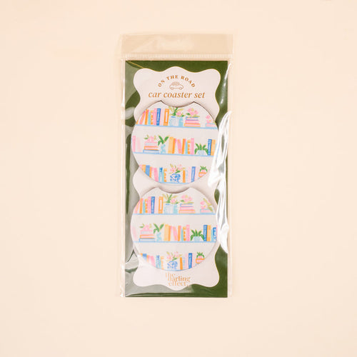 The Book Shelfie On the Road Car Coaster Set by The Darling Effect includes two absorbent coasters for your car cup holder, featuring vibrant books and plants on shelves. Packaged in clear plastic with branded labeling.