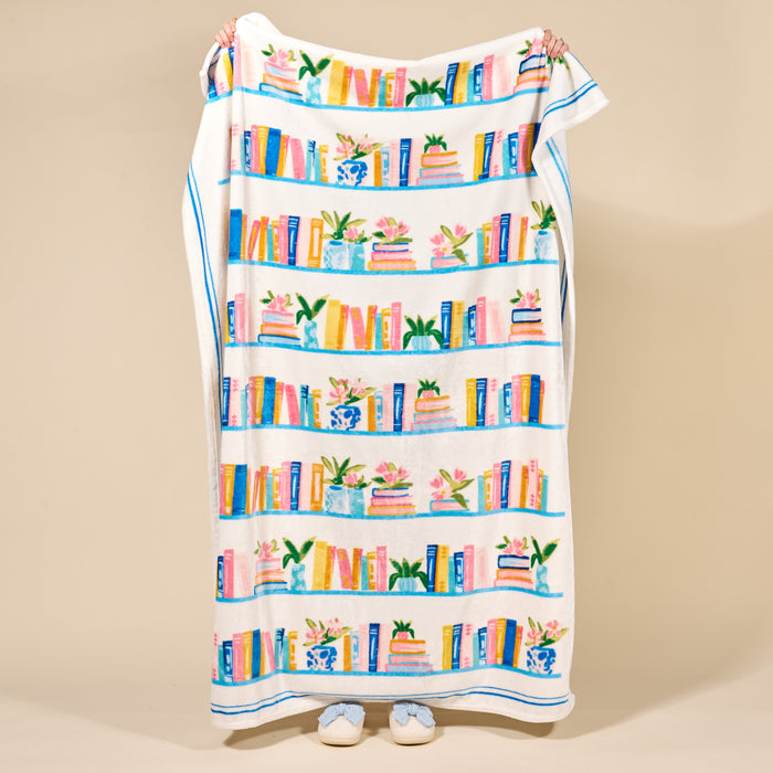 A person holds up The Darling Effect's Book Shelfie Snuggle Up Plush Blanket, showing off its cozy design featuring colorful books, plants, and vases on shelves. Only their hands and slippered feet are visible against a beige background.