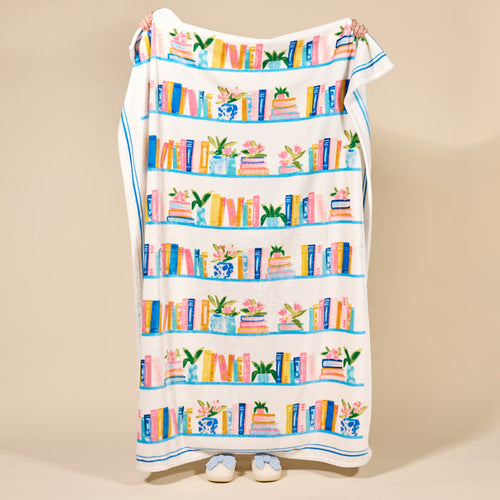 A person holds up The Darling Effect's Book Shelfie Snuggle Up Plush Blanket, showing off its cozy design featuring colorful books, plants, and vases on shelves. Only their hands and slippered feet are visible against a beige background.