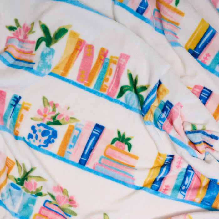 The Darling Effect's Book Shelfie Snuggle Up Plush Blanket features colorful illustrations of books, plants, and vases on blue shelves with pink, yellow, blue, and green accents for a cozy, playful touch to any space.