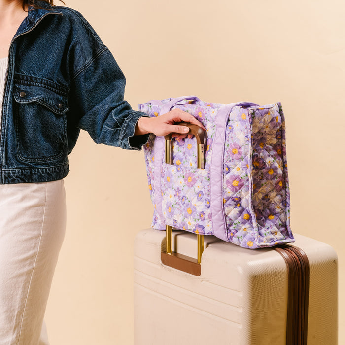 A person in a denim jacket and white dress stands next to a beige suitcase, holding the-darling-effect’s Bouquet Beauty Purple The Everyday Quilted Tote Bag by its handles, against a light beige background.