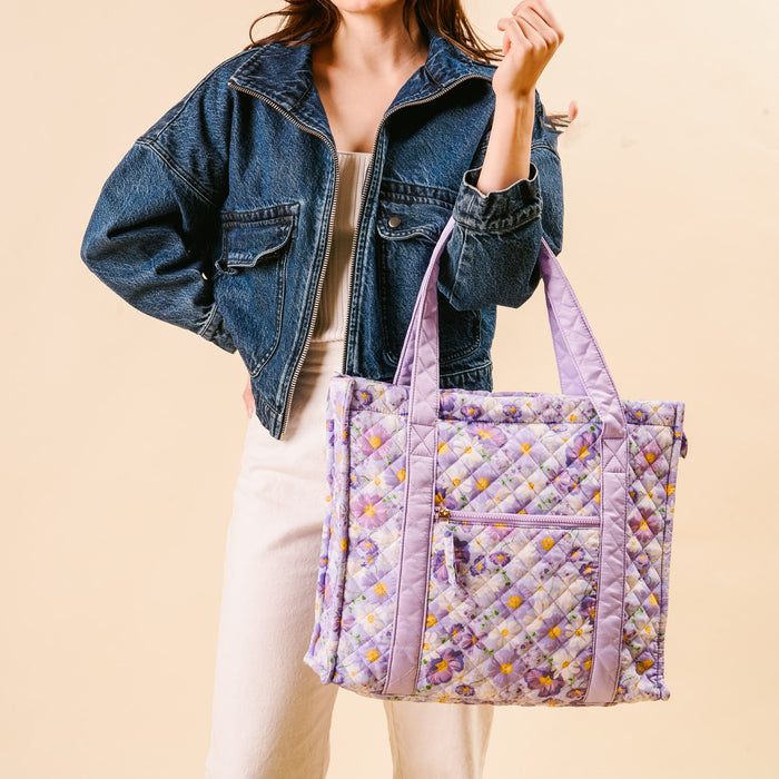 A woman in a denim jacket and white pants holds the-darling-effect’s Bouquet Beauty Purple The Everyday Quilted Tote Bag, featuring purple handles and a pastel floral pattern on a beige background.