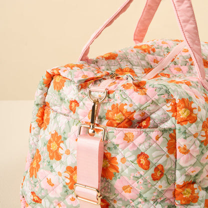 A close-up of the-darling-effect’s Bouquet Beauty Sage On-the-Go Quilted Weekender Bag, featuring a pink, orange, and white floral design with matching handles and detachable strap with gold hardware, resting on a light-colored surface.