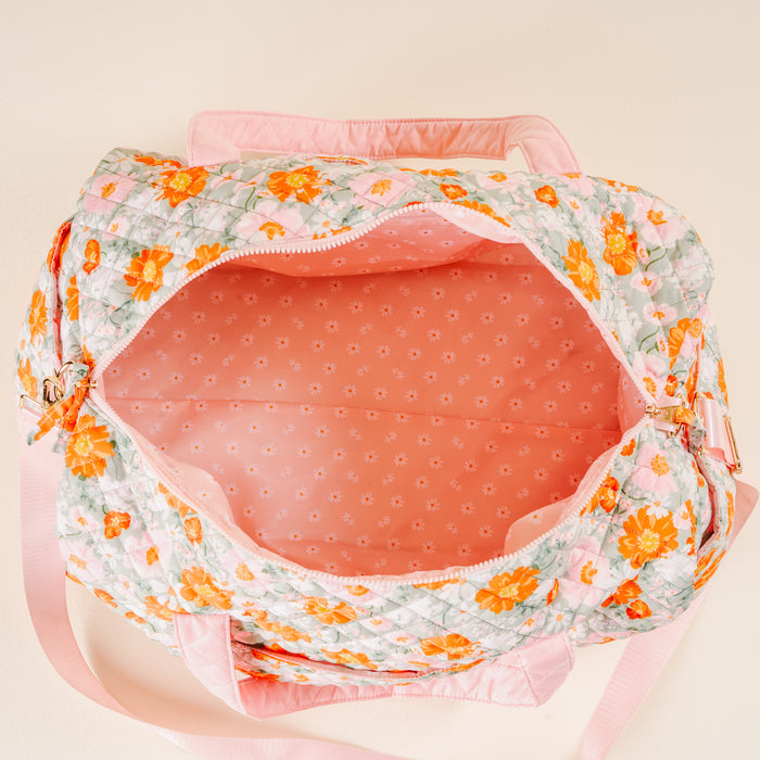 The Bouquet Beauty Sage On-the-Go Quilted Weekender Bag by the-darling-effect features a colorful floral print, pink handles, and an adjustable shoulder strap, shown open against a light peach background.