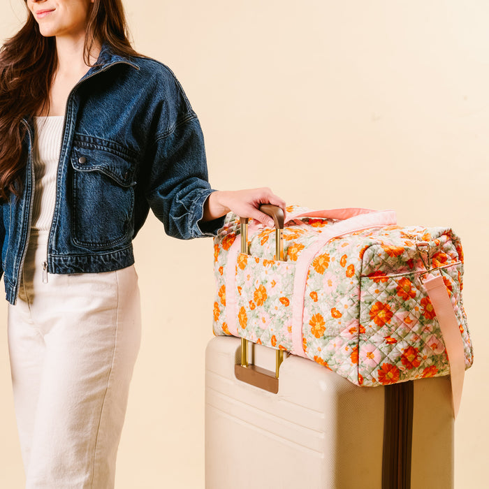 A woman in a denim jacket and white pants stands beside beige luggage topped with the-darling-effect’s Bouquet Beauty Sage On-the-Go Quilted Weekender Bag, holding its handle against a light, neutral background.