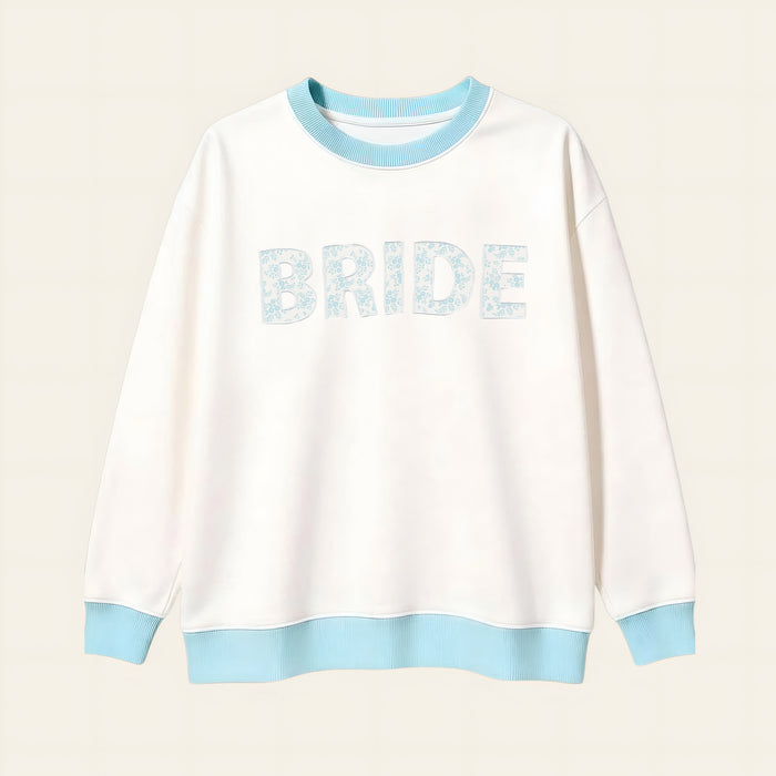The Darling Effect Bride Oversized Crew is a cozy white sweatshirt with a light blue collar, cuffs, and hem. "BRIDE" appears in large light blue floral letters on a soft cream grid background.